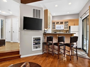 A modern kitchen with a flat-screen TV, wooden cabinets, and a bar area with stools.