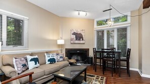 A living room with a beige sofa, a fireplace, a dining table with chairs, and large windows with blinds. - Three Ridges 84 with Wintergreen Advantage (Wintergreen)