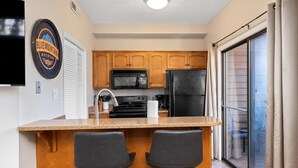 A kitchen with wooden cabinets, a black refrigerator, a microwave, and a coffee maker. There is a bar area with two chairs and a countertop.