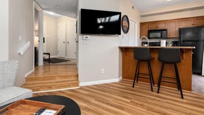 A modern kitchen with wooden cabinets, a black refrigerator, and a microwave. There is a bar area with two stools and a television mounted on the wall.