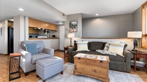 Een moderne woonkamer met een grijze bank, een grijze fauteuil en een houten salontafel. Er is een keukenhoek met houten kastjes en een koelkast op de achtergrond.