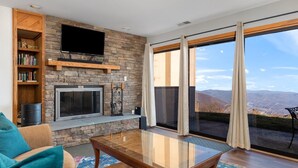 A living room with a stone fireplace, a flat-screen TV, a bookshelf, and a large sliding glass door leading to a balcony with a mountain view. - Cliffs 21 with Wintergreen Advantage (Wintergreen)