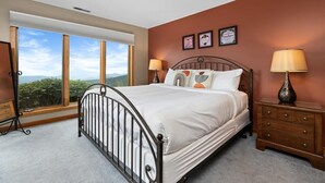 A bedroom with a large window, a bed with white bedding, two bedside tables with lamps, and framed certificates on the wall.