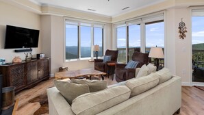 A spacious living room with a large sofa, a coffee table, and a TV mounted on the wall. There are large windows offering a view of the outdoors.