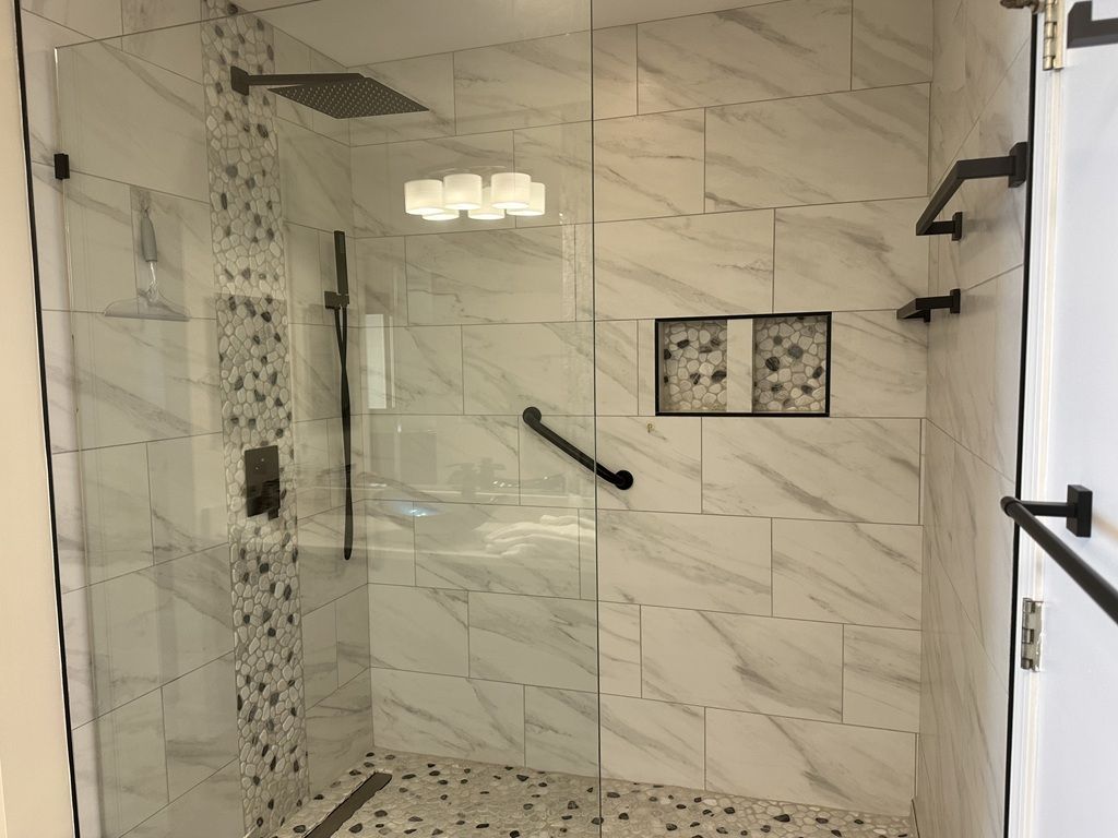 Un espace douche clos par des parois de verre, doté d’un sol en marbre et de carreaux muraux, d’un pommeau de douche à main et d’un luminaire fixé au mur.