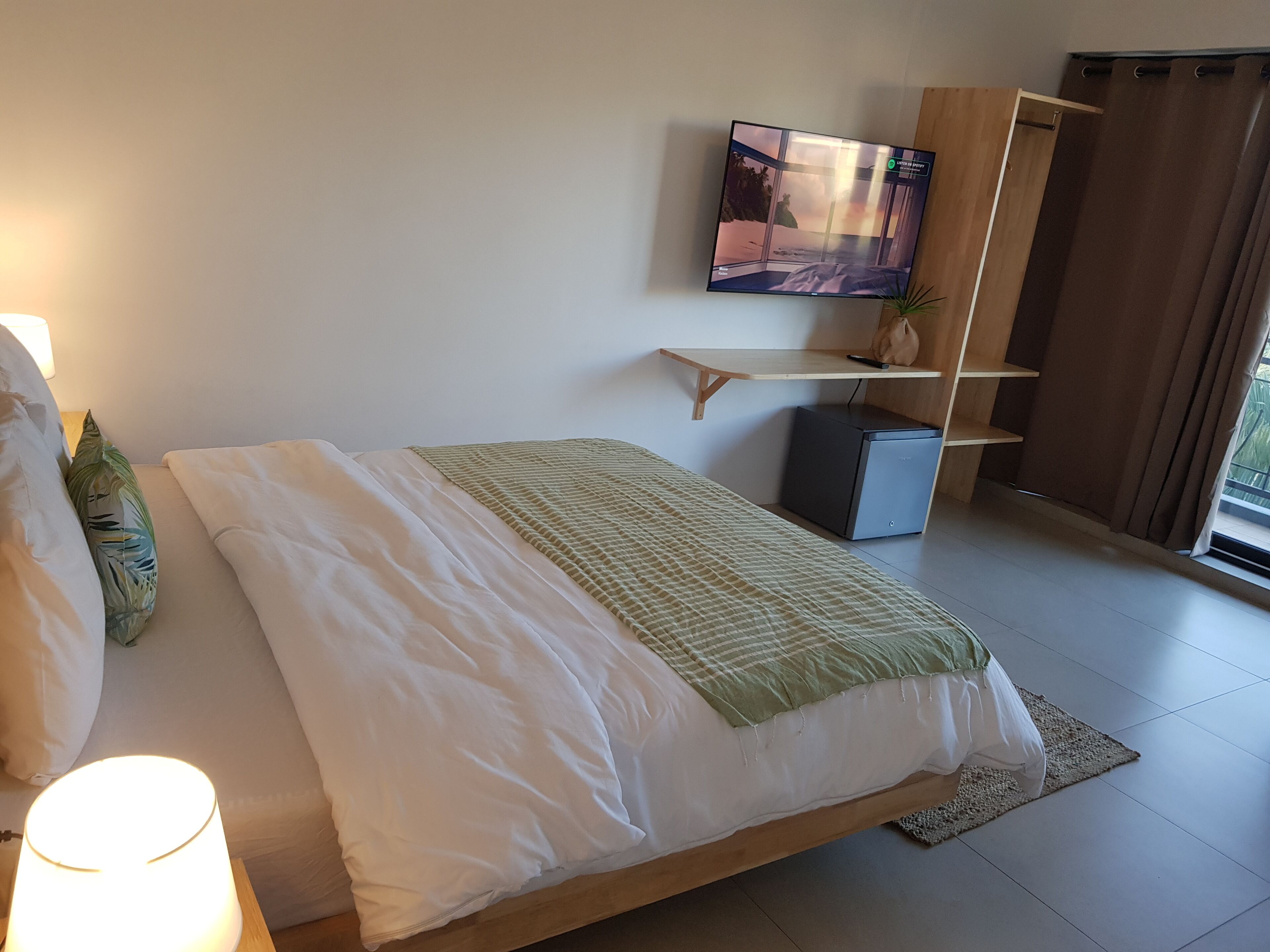 A bedroom with a bed, a TV mounted on the wall, a small desk, and a window with curtains.