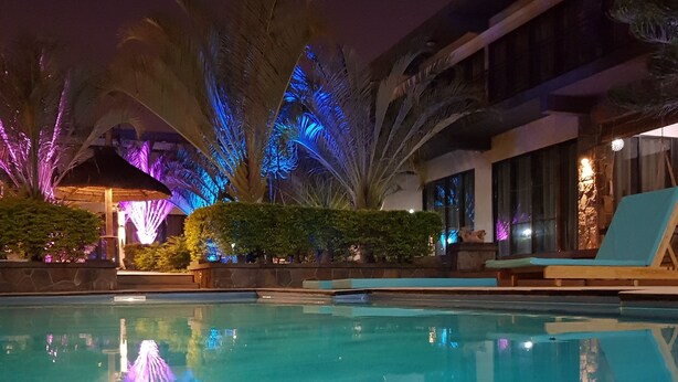 A swimming pool with a water fountain, surrounded by palm trees and a building with a balcony. - Villas Rocher (Fond du Sac)
