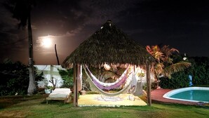 Una cabaña con techo de paja, una hamaca, una cama pequeña y una piscina en un entorno tropical.