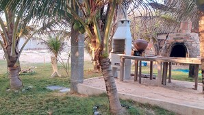 Un patio al aire libre con un horno de piedra, mesas y sillas de madera, rodeado de palmeras y suelo arenoso.
