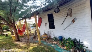 Un edificio rústico con fachada blanca, puerta de madera y una ventana pequeña. Hay zonas de estar al aire libre con bancos y una hamaca.