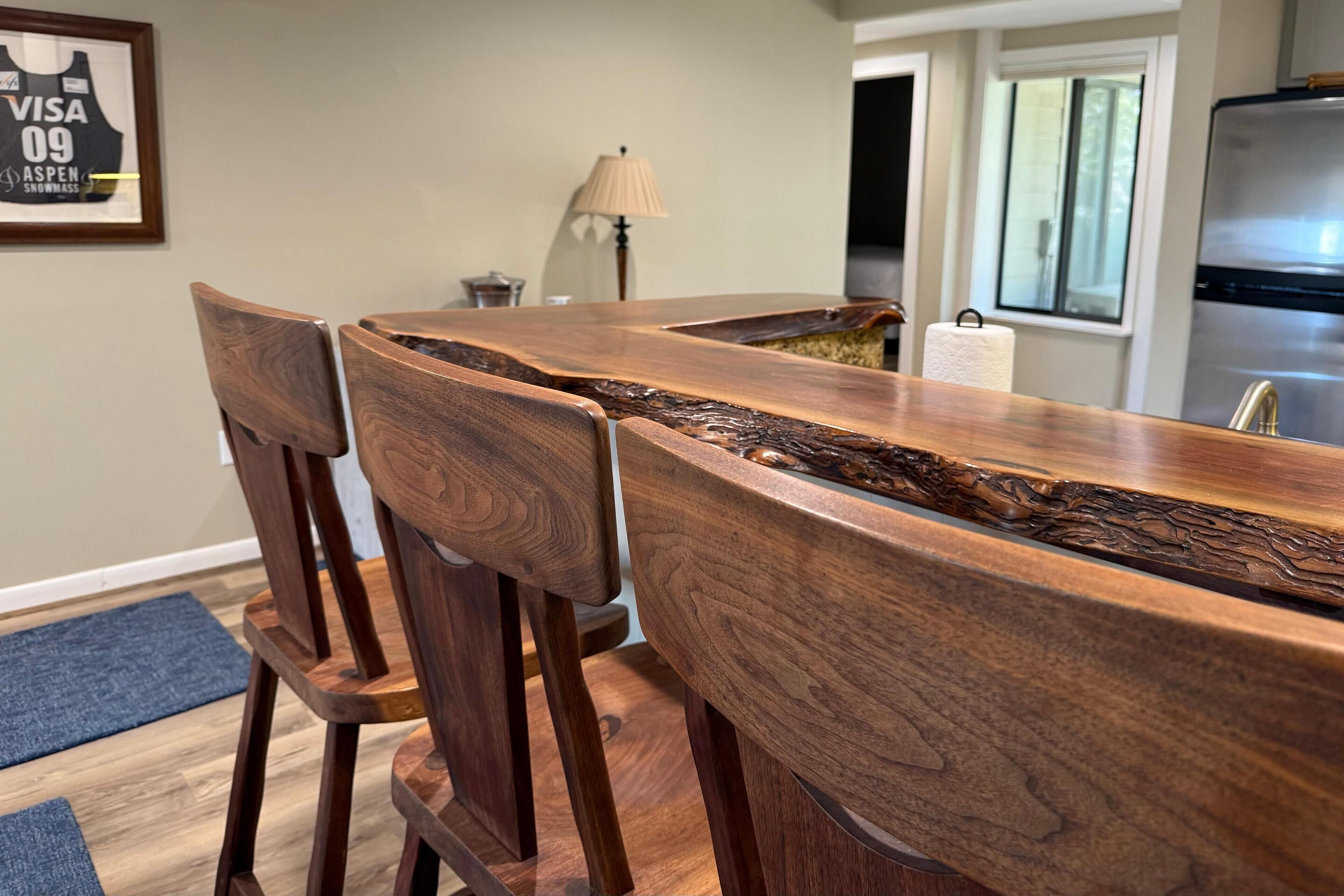 A wooden dining table with a unique design, surrounded by wooden chairs. A lamp and a refrigerator are visible in the background.