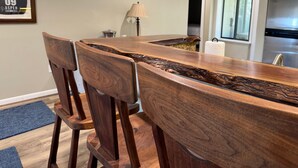 A wooden dining table with a unique design, surrounded by wooden chairs. A lamp and a refrigerator are visible in the background.