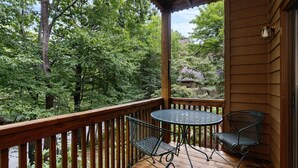 Une terrasse en bois avec une table ronde et deux chaises, entourée d’une balustrade en bois, donnant sur une zone boisée.