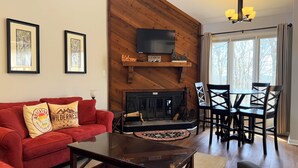 Una sala de estar con un sofá rojo, una chimenea, una mesa de comedor con sillas, un televisor montado en una pared de madera y cuadros enmarcados en la pared.