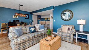 Un salón con una pared azul, dos sofás estampados, una mesa de centro y un comedor con una cocina al fondo.