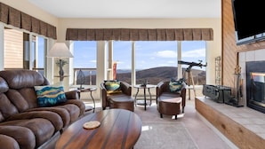 Una sala de estar moderna con sofás de cuero, una mesa de centro de madera y una chimenea. Ventanas amplias ofrecen vistas a las montañas.