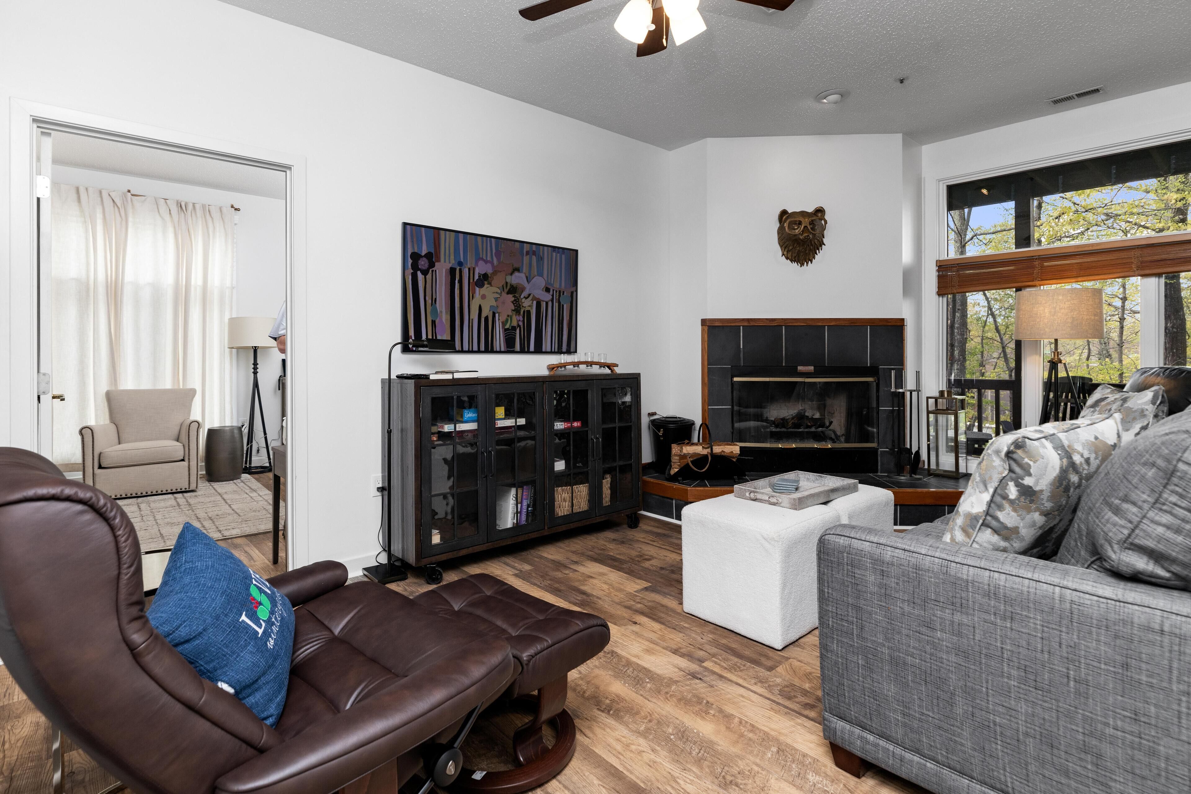A living room with a leather chair, a grey sofa, a fireplace, a bookshelf, and a large window.