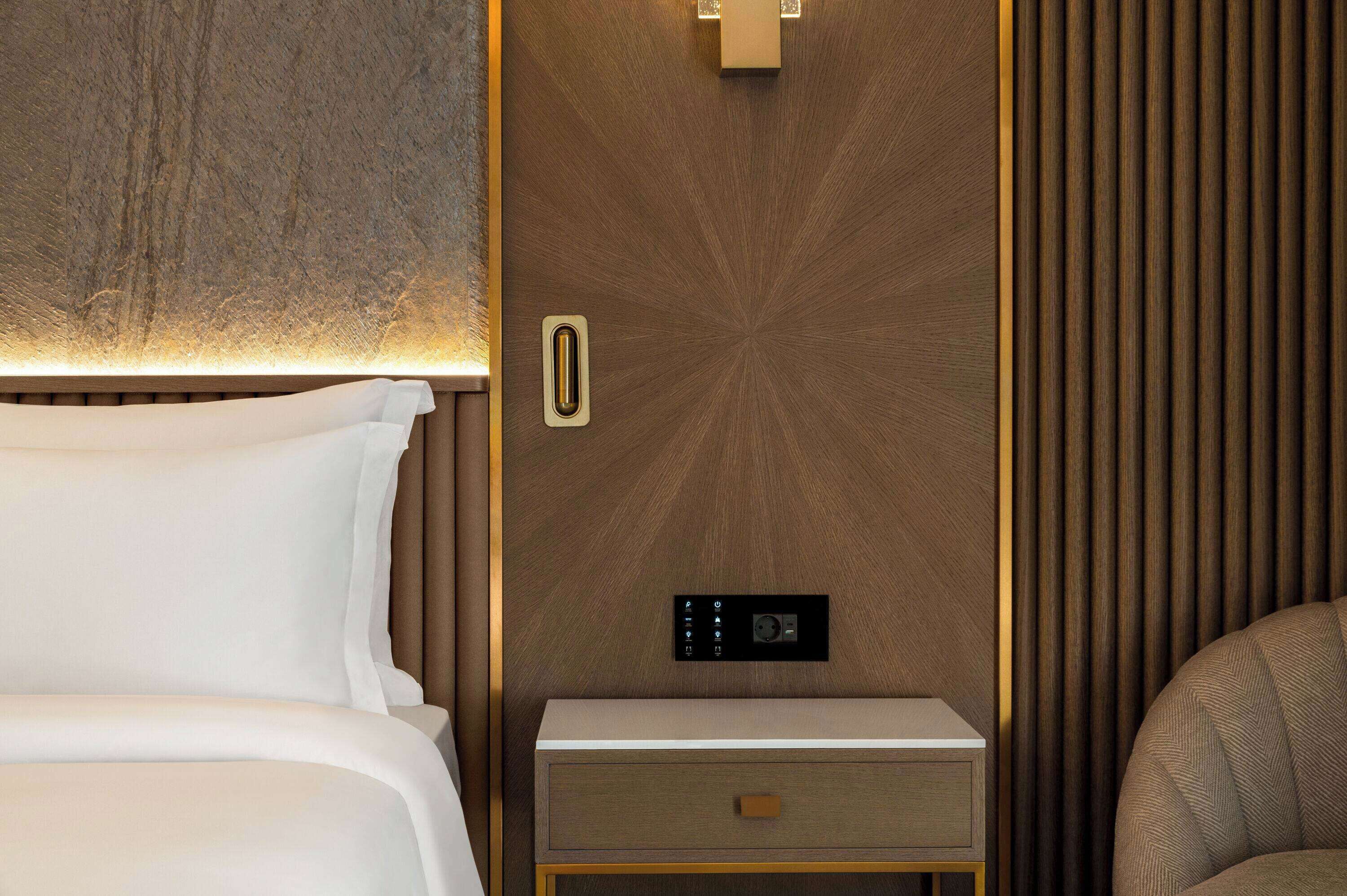 A modern hotel room with a bed, bedside table, and a control panel on the wall.