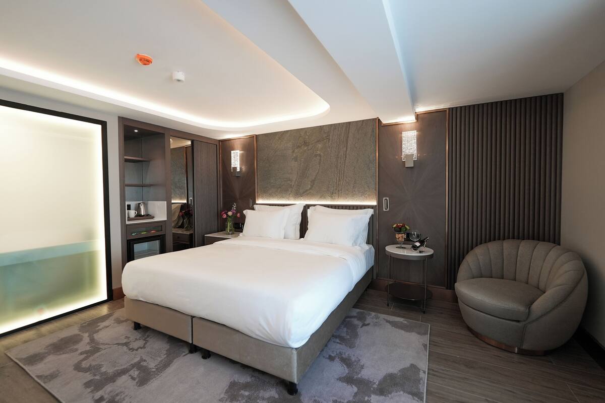 Ein modernes Hotelzimmer mit einem großen Bett, einem runden Sessel, einem Nachttisch mit Lampe und einem eingebauten Kleiderschrank.