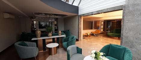 Un interior moderno con techos altos, una zona de estar central con sofás verdes, una mesa de centro blanca y una cocina a la vista.