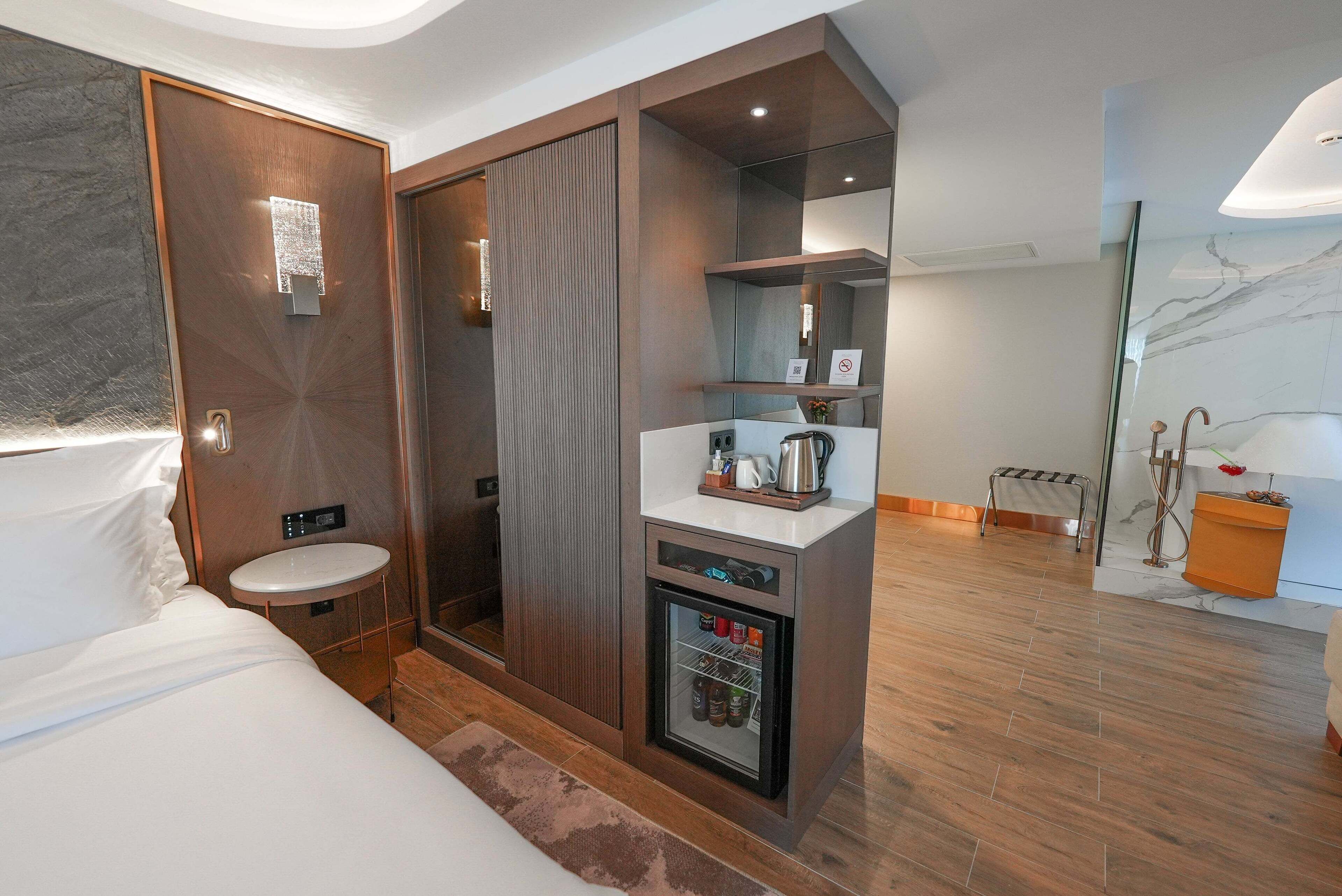 A modern hotel room with a bed, a small bathroom, a minibar, and a shower area.