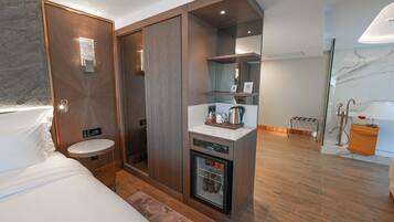 A modern hotel room with a bed, a small bathroom, a minibar, and a shower area.