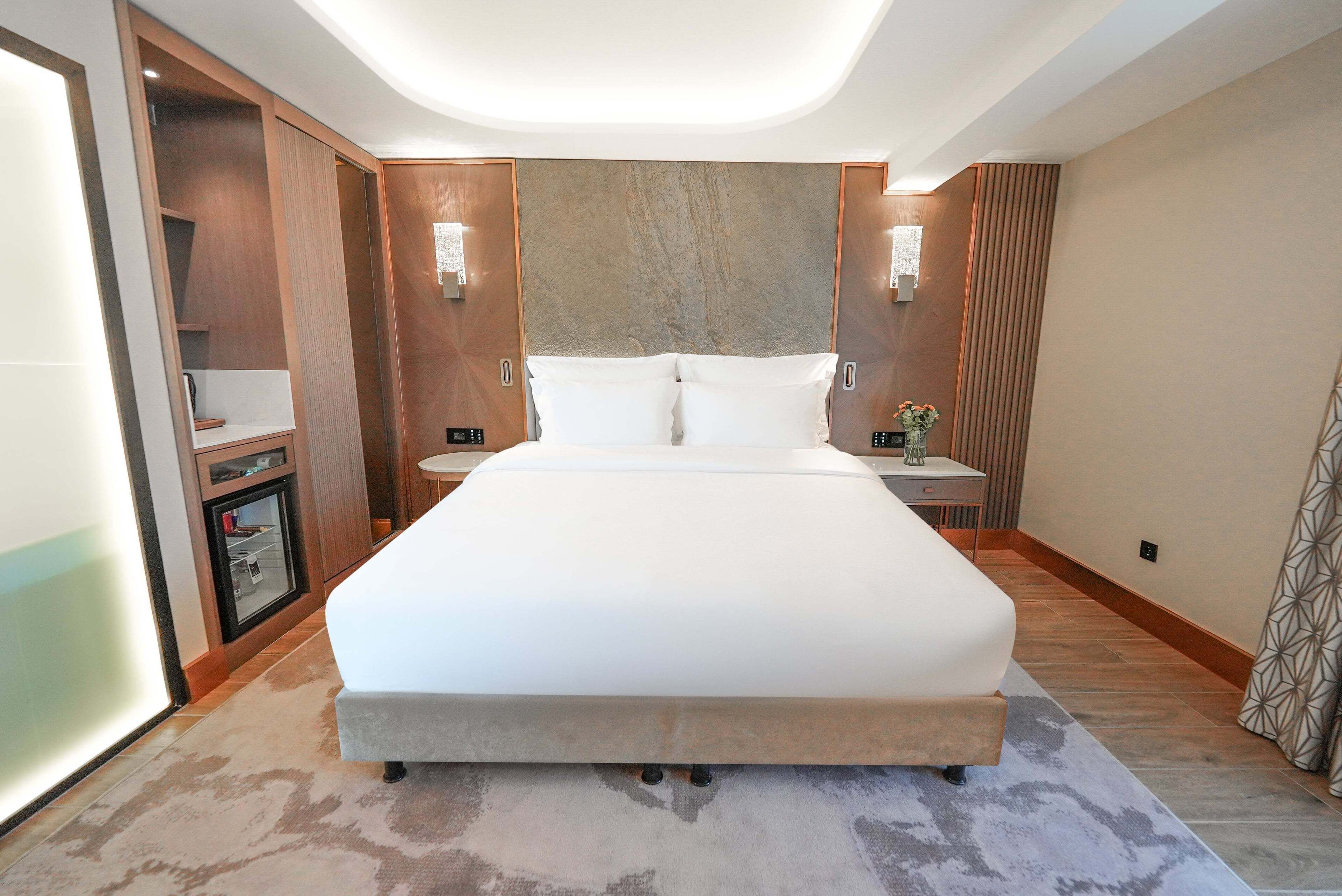 A modern hotel room with a large bed, built-in wardrobe, bedside tables, and a small desk with a lamp.