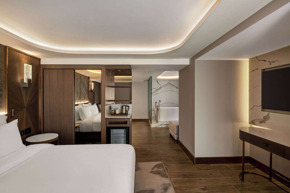 Ein modernes Hotelzimmer mit einem großen Bett, einem kleinen runden Tisch, einem Schreibtisch, einem Badezimmer mit Badewanne und einem an der Wand befestigten Fernseher.