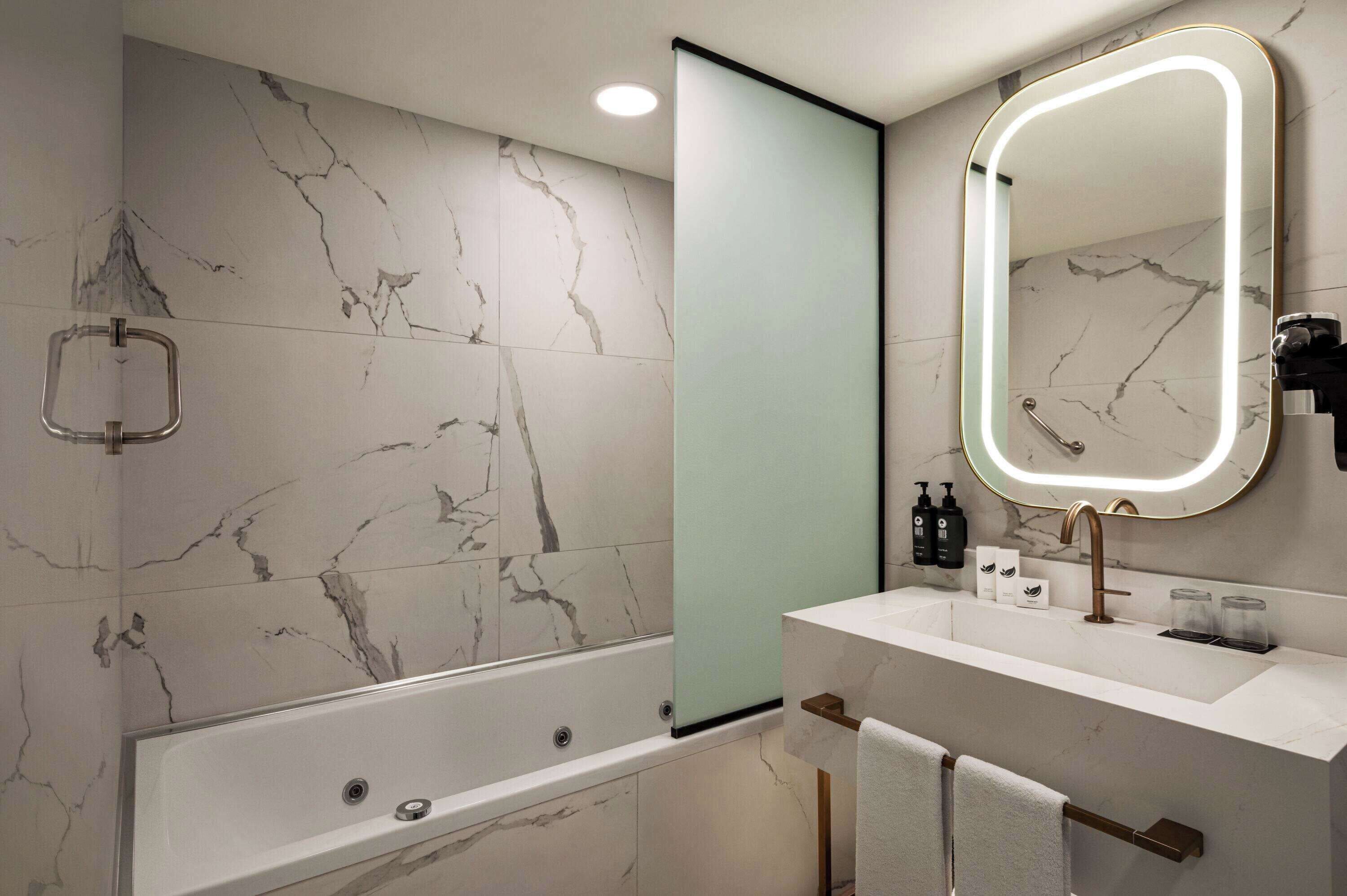 Une salle de bain moderne avec un mur recouvert de carreaux de marbre, une baignoire, un lavabo avec un miroir et des articles de toilette posés sur le comptoir.
