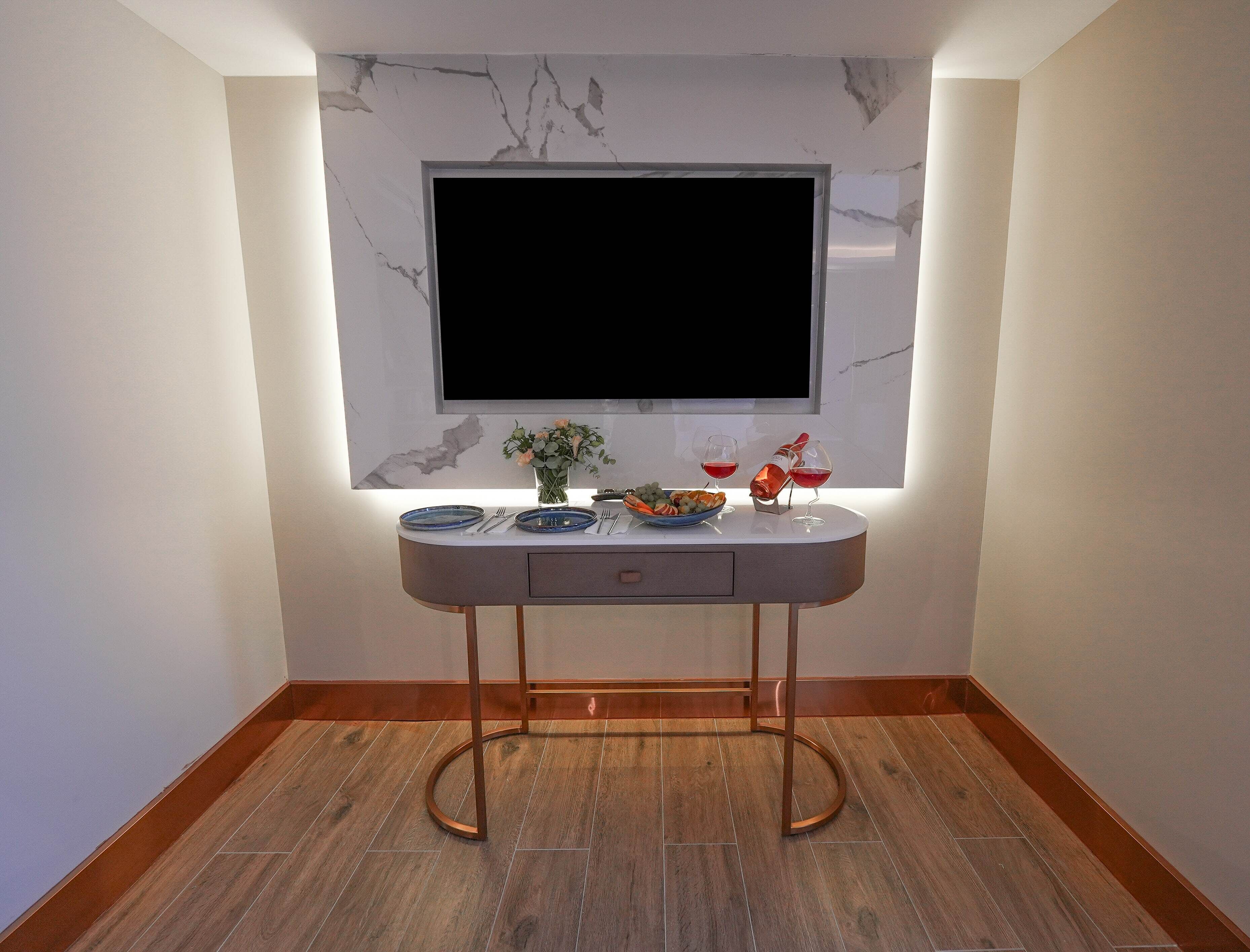 A modern living room with a flat-screen TV mounted on a marble wall, a dining table with plates, cutlery, and a vase of flowers.