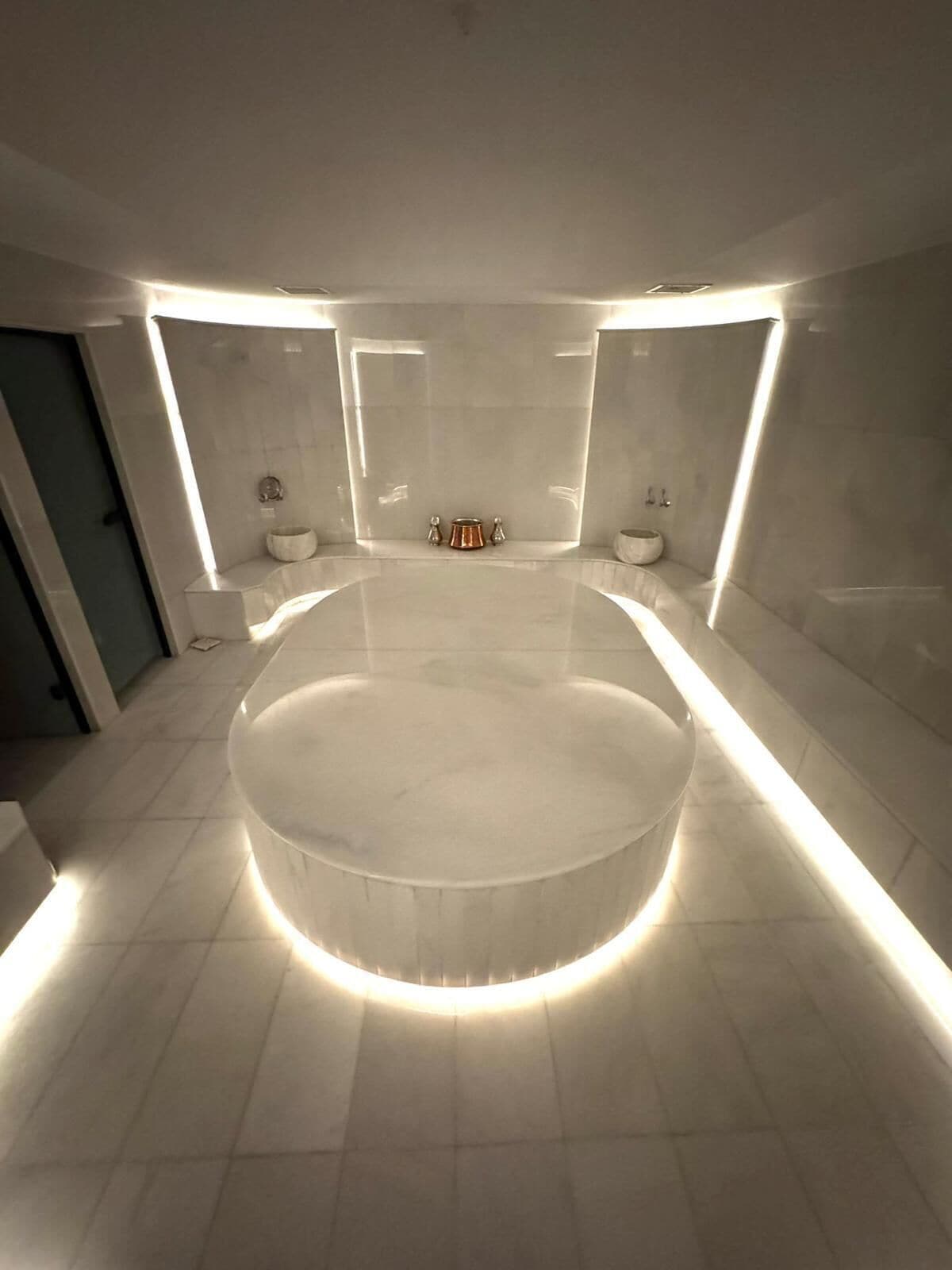 A modern bathroom with a central circular bathtub, illuminated by underfloor lighting, and flanked by two sinks with matching fixtures.