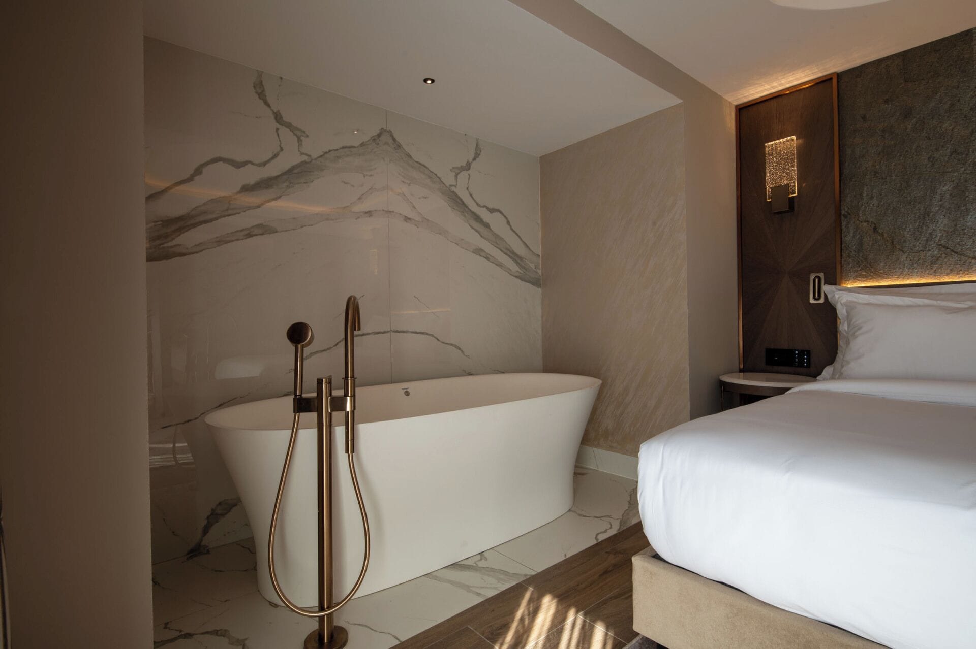A modern bathroom with a freestanding bathtub, a shower with a handheld showerhead, and a bedside table with a lamp.