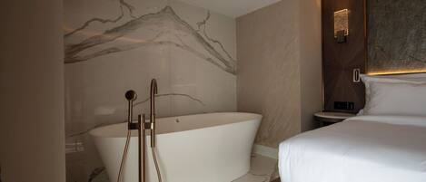 A modern bathroom with a freestanding bathtub, a shower with a handheld showerhead, and a bedside table with a lamp.