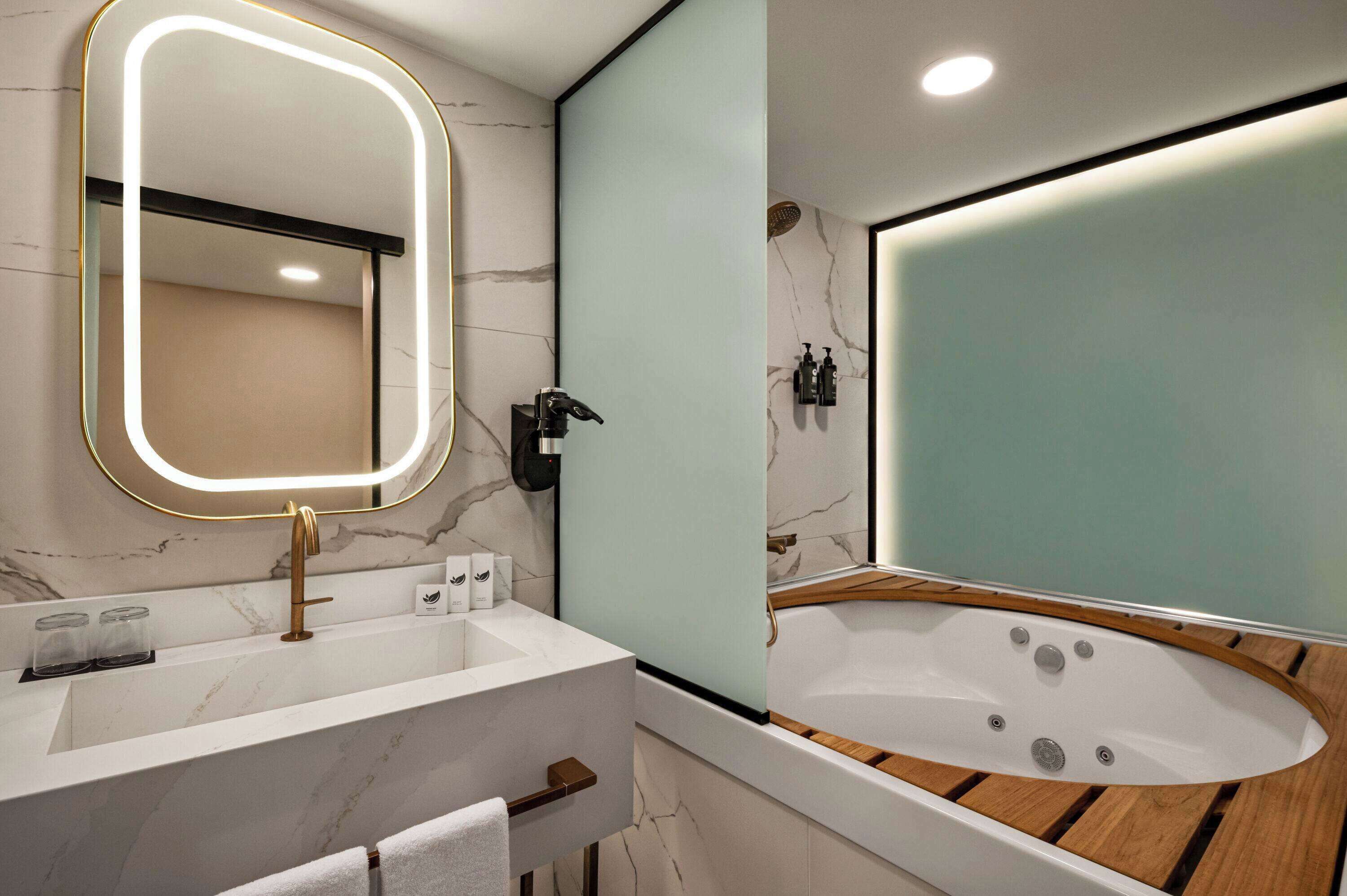 Un baño moderno con lavamanos de mármol, un espejo grande con iluminación LED, una bañera independiente y un mueble de tocador de madera.