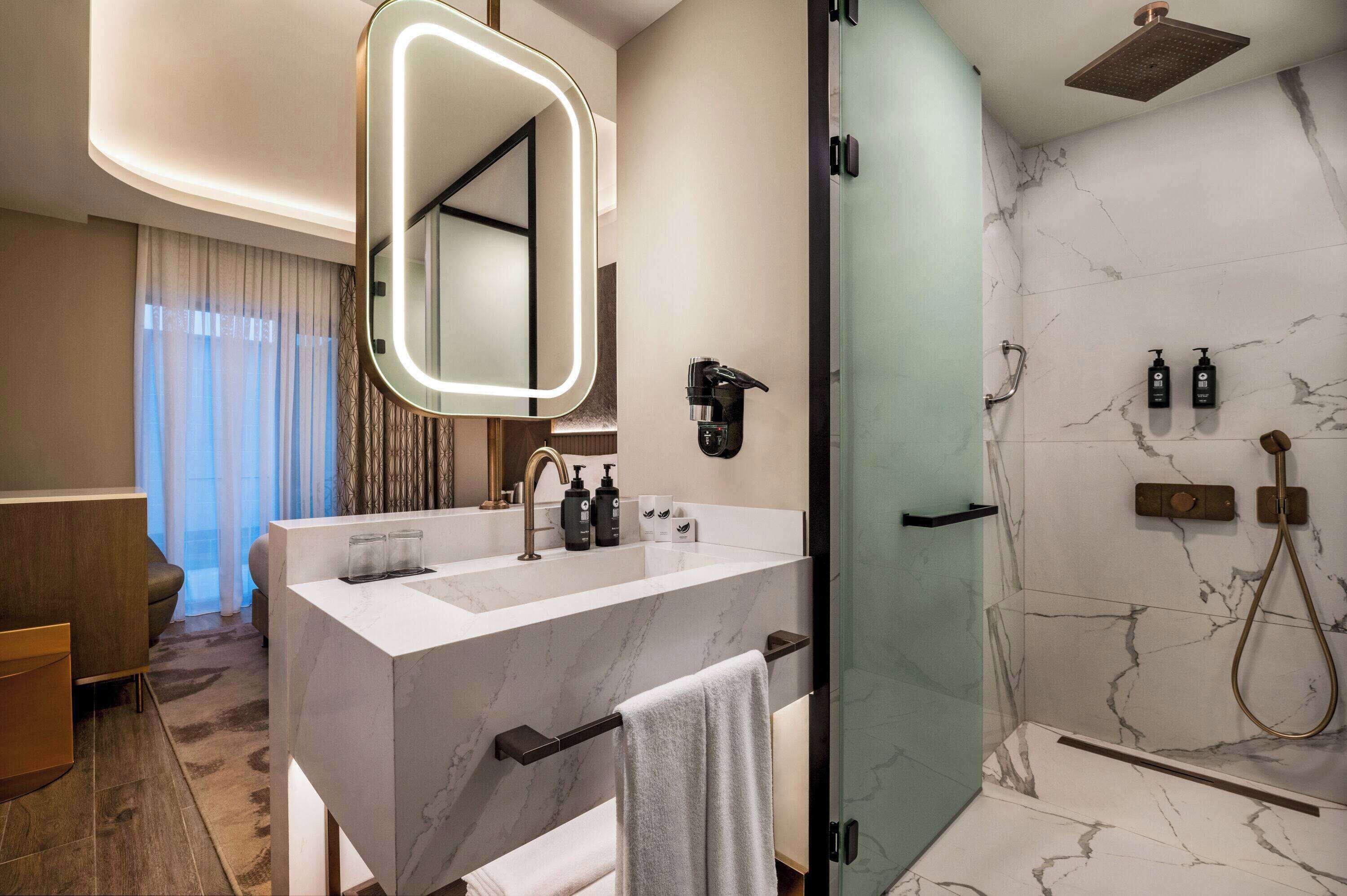A modern bathroom with a large mirror, dual sinks, a shower with a handheld showerhead, and marble walls.