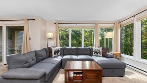 A modern living room with a sectional sofa, a wooden coffee table, and large windows offering a view of the outdoors.
