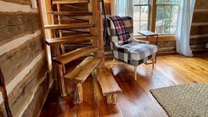 A log cabin interior with a wooden spiral staircase, a plaid armchair, and a window with curtains.
