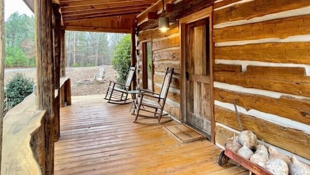 A log cabin with a wooden porch, two rocking chairs, and a wheelbarrow with stones.