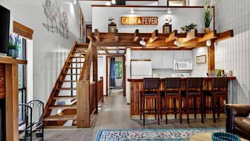 Interior de una cabaña con escalera de madera, cocina con armarios blancos y una zona de bar.
