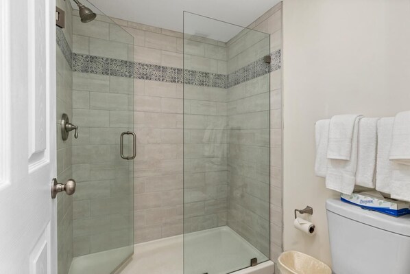 A glass-enclosed shower with a tiled floor and a white toilet with a blue box on top.