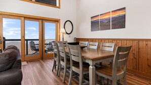 Un comedor con mesa y sillas de madera, vista al exterior a través de puertas de vidrio, y una pared con tres cuadros de paisajes.