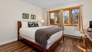 Un dormitorio con una cama de madera, una ventana con cortinas, una mesita de noche con una lámpara y dos cuadros en la pared.