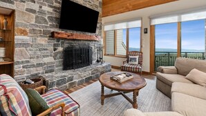 A stone fireplace with a flat-screen TV, a cushioned sofa, a round coffee table, and a view of the outdoors.