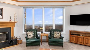 A living room with a fireplace, two green armchairs with animal-themed cushions, a wooden coffee table, a TV on the wall, and a large window with a view of the outdoors. - Highridge Court 19 with Wintergreen Advantage (Wintergreen)