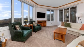 A spacious living room with large windows, a flat-screen TV, and a variety of seating options including a green leather sofa, a beige sofa, and a wooden coffee table. - Highridge Court 19 with Wintergreen Advantage (Wintergreen)