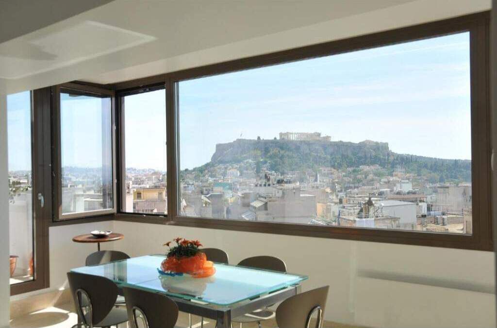 Une chambre moderne dotée d’une grande fenêtre donnant sur le paysage urbain, d’une table en verre avec des chaises et d’une petite table ronde surmontée d’un vase.