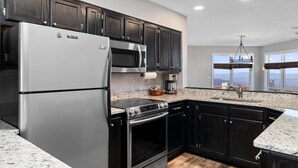 Cozinha moderna com armários escuros, eletrodomésticos de aço inoxidável, bancadas de granito e vista através de amplas janelas.