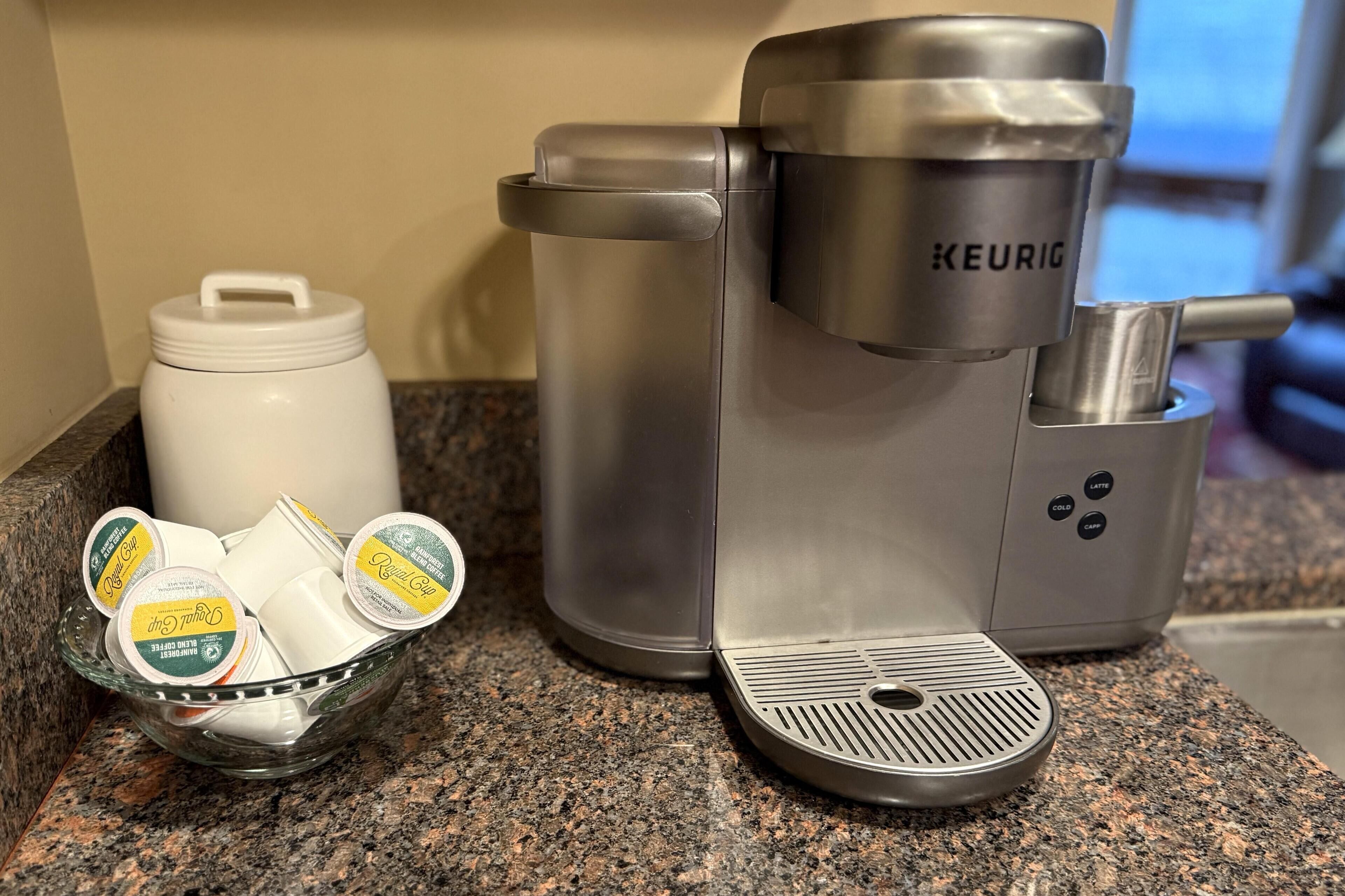 A Keurig coffee maker on a granite countertop with a coffee pod holder and a white container.