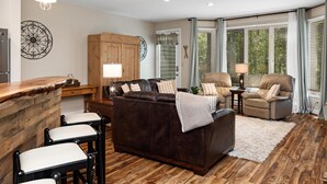 Ein gemütliches Wohnzimmer mit einer dunkelbraunen Ledersofa, zwei beigen Sesseln und einem Holztisch. Ein weißer Teppich bedeckt den Holzboden, und eine Holzkommode steht an der Wand.