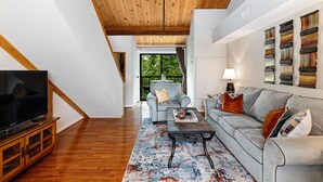 Un salon moderne avec un plafond en bois, un canapé gris, une table basse et un téléviseur posé sur un meuble en bois.